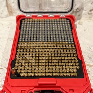 May include: A red plastic ammunition storage case with a clear lid, filled with numerous brass-colored bullets. The bullets are neatly arranged in rows within a black insert. The case is designed for secure storage and transport of ammunition.