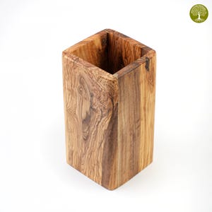 Puede incluir: Un recipiente cuadrado de madera con un patrón de vetas de madera natural. El recipiente está hecho de madera de olivo y tiene forma rectangular. El interior es hueco y el exterior tiene un acabado liso. Ideal para guardar bolígrafos, lápices u otros objetos pequeños.