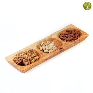Può includere: Un vassoio di legno con tre scomparti, ciascuno riempito con frutta secca diversa. Il vassoio contiene noci, pistacchi e mandorle. Il legno ha una venatura naturale e un colore marrone chiaro caldo. Ideale per servire snack.