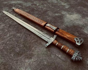 May include: A double-edged steel sword with a patterned blade, a wooden grip featuring silver-colored metal bands, and a matching pommel. Beside it lies a wooden scabbard decorated with intricate carved knotwork and silver-colored metal accents.
