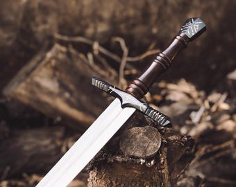 May include: A metal sword featuring a polished silver blade and a dark brown wooden handle. The crossguard and pommel are black with intricate white engraved markings. The sword is positioned at an angle with the blade extending outward.