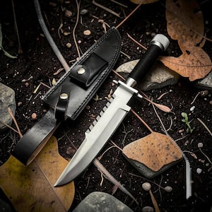 May include: A survival knife with a serrated blade and black handle rests beside its black leather sheath. The knife is silver and black, with a textured handle and a guard. The sheath has a belt loop and a snap closure.