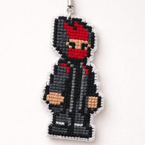 May include: A pixelated charm featuring a figure in a black hooded jacket, red mask, and tan skin. The charm is attached to a black cord and silver-toned metal clasp. The figure is made of a cross-stitch design.