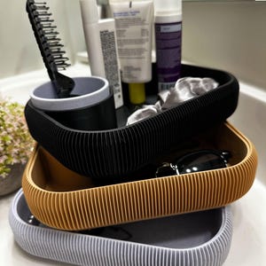 May include: Three stacked, ribbed trays in black, gold, and gray, holding toiletries and accessories. A black comb stands in a matching cup. Sunglasses and various cosmetic products are visible, suggesting bathroom organization.
