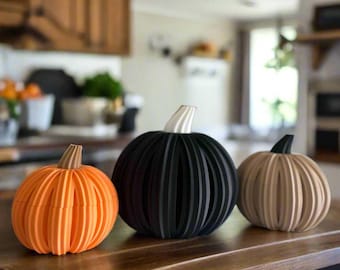 May include: Three decorative pumpkins in orange, black, and tan, with ribbed designs. The pumpkins have stems in contrasting colors. The pumpkins are arranged on a wooden surface, perfect for fall decor.
