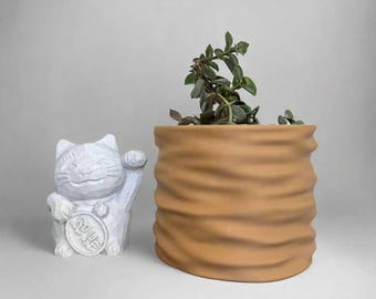 May include: A white ceramic lucky cat figurine with a raised paw and a coin in its paw, next to a brown ceramic planter with a green plant inside.