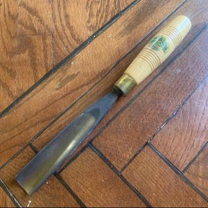 Può includere: Uno scalpello per la lavorazione del legno con un manico in legno chiaro e una lama in metallo. L'impugnatura ha una forma cilindrica con scanalature e una ghiera in ottone. La lama è scura e angolata, progettata per tagli precisi. Il manico ha il testo "Fois".