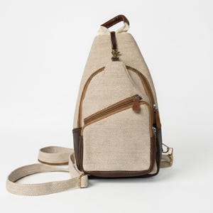 May include: A beige and brown sling backpack featuring a herringbone pattern on the main body. The bag includes a diagonal zippered front pocket, brown leather-like trim, a top handle, and an adjustable woven strap with metal hardware.