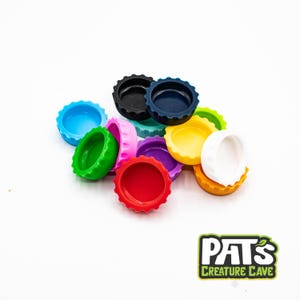May include: A collection of colorful bottle caps in various shades, including blue, green, red, and yellow. The caps are arranged on a white background. The logo for Pat's Creature Cave is in the bottom right corner.