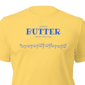 May include: A yellow t-shirt with a blue graphic of a stick of butter with the text "GRADE AA BUTTER NET WT: 4 OZ. (113g)" and a measurement scale showing tablespoons.