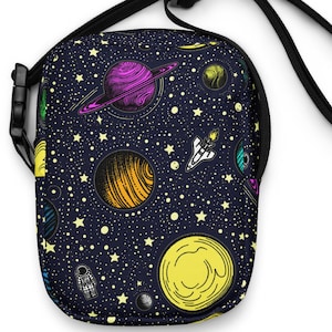 May include: A black crossbody bag with a colorful space-themed pattern. The design features planets, stars, and a rocket ship against a dark blue background. The bag has a black adjustable strap and a black buckle.