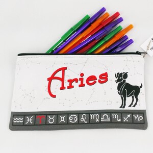 May include: White pencil case with the word "Aries" in red, a black ram illustration, and various zodiac symbols. The case is filled with colorful pens. A tag with the words "Firebolt Creations" is attached.