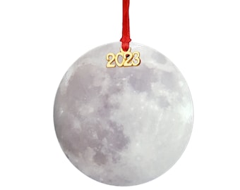 May include: A round, white, ceramic ornament with a picture of the moon. The ornament has a red ribbon and a gold charm with the year 2023 on it.