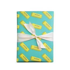 May include: A wrapped gift with teal wrapping paper featuring a repeating pattern of yellow butter sticks. The gift is tied with a cream-colored satin ribbon in a bow. The word "BUTTER" is printed on the butter sticks.