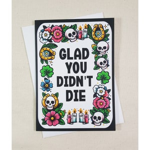 May include: A black and white greeting card with a white background and black border. The card features a colorful illustration of skulls, flowers, clovers, horseshoes, and candles. The text on the card reads "Glad you didn't die."