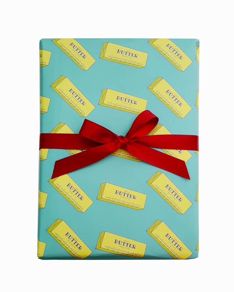 May include: A wrapped gift with a teal background featuring a repeating pattern of yellow butter sticks with the word "BUTTER" printed on them. The gift is tied with a red ribbon and bow.