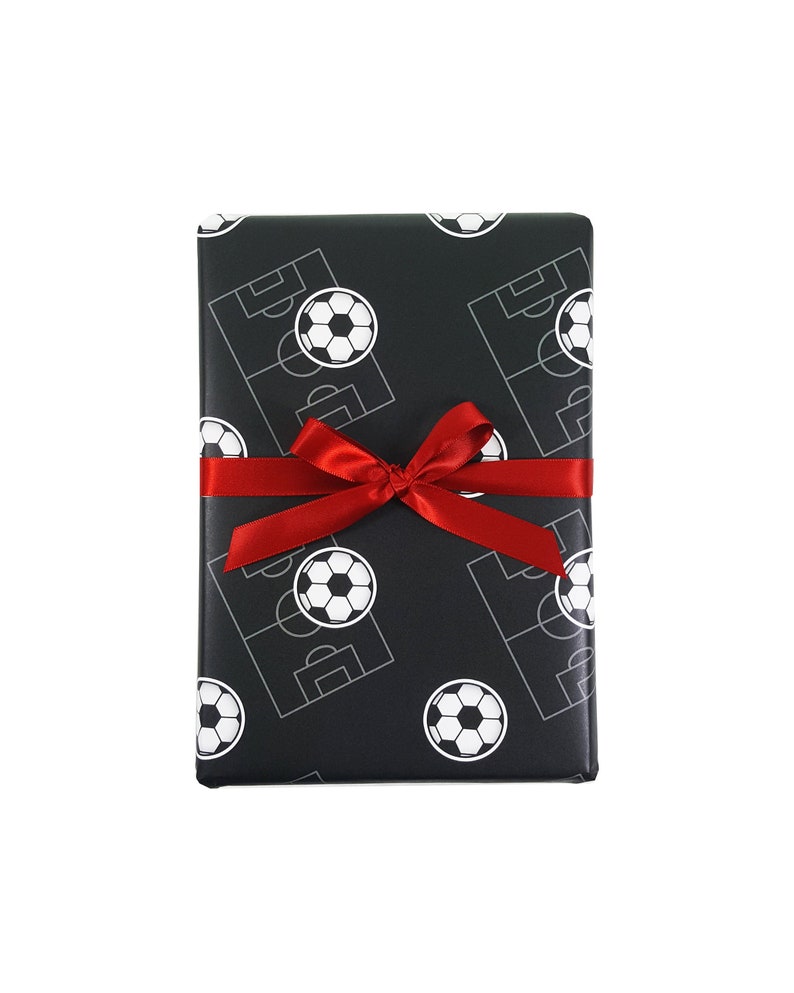 Puede incluir: Una caja de regalo negra con una cinta roja. La caja est&aacute; envuelta en papel con un patr&oacute;n de balones de f&uacute;tbol blancos y campos de f&uacute;tbol.