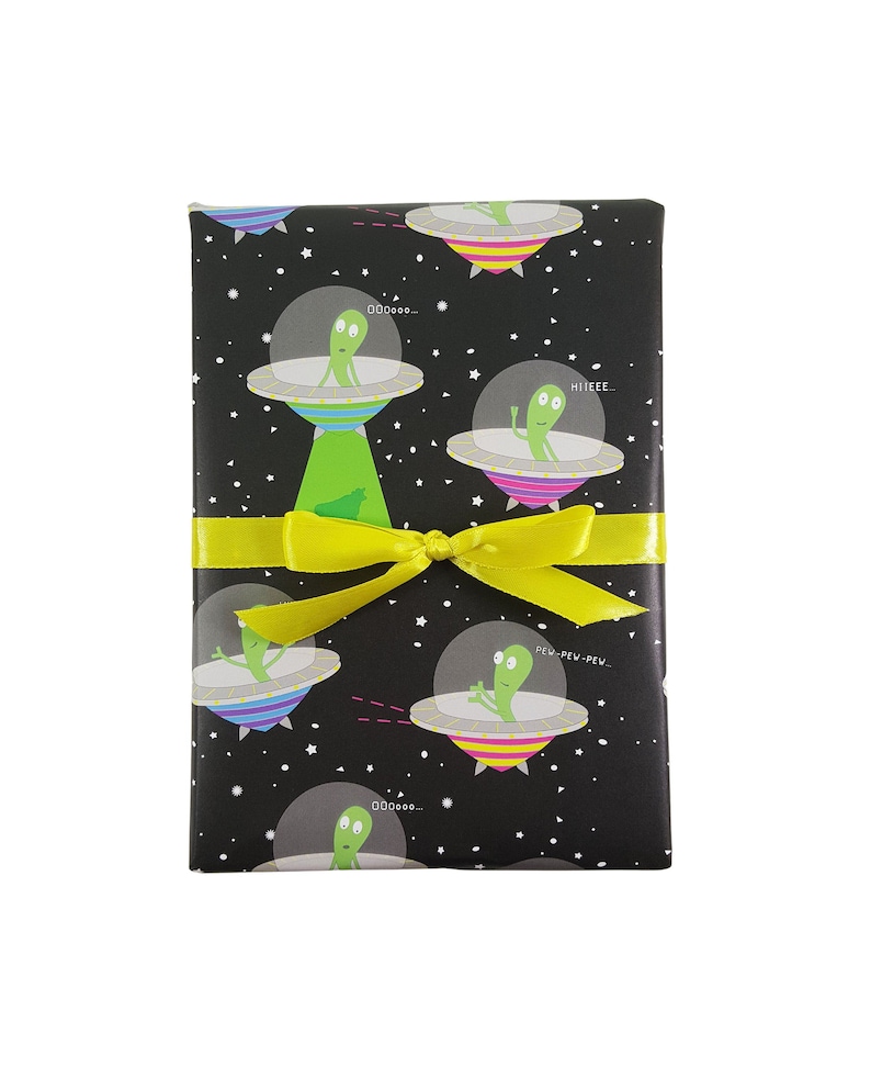 May include: Black gift wrap with a repeating pattern of green aliens in flying saucers on a black background with white stars. The aliens have different expressions and some have speech bubbles with text like "pew-pew-pew" and "hieee". A yellow ribbon is tied around the gift wrap.