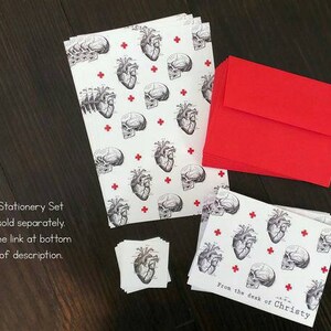 May include: A stationery set featuring a black and white anatomical heart and brain pattern on white paper with red crosses. The set includes note cards, envelopes, and small stickers. The text "From the desk of Christy" is printed on the note cards.