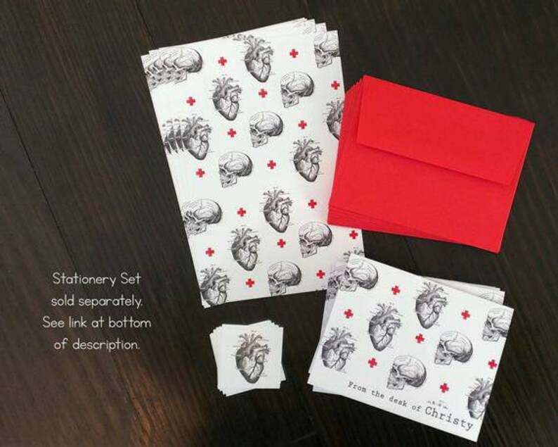May include: A stationery set featuring a black and white anatomical heart and brain pattern on white paper with red crosses. The set includes note cards, envelopes, and small stickers. The text "From the desk of Christy" is printed on the note cards.