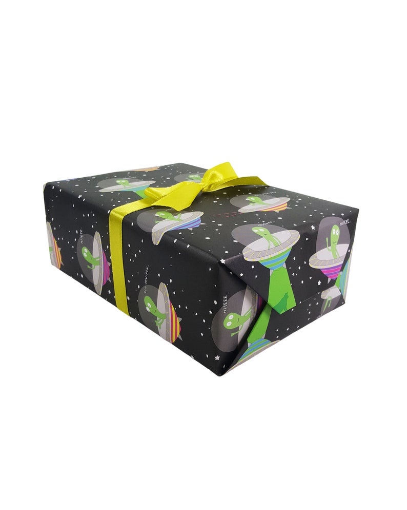 May include: A black gift box with a yellow ribbon. The box is wrapped in paper with a pattern of green aliens in flying saucers against a black background with white stars.