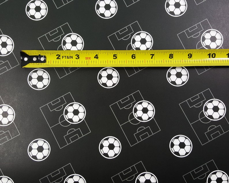 Puede incluir: Una cinta m&eacute;trica amarilla con marcas negras se coloca sobre un fondo negro con un patr&oacute;n repetido de balones de f&uacute;tbol blancos y campos de f&uacute;tbol. La cinta indica "2 FT & IN 3 4 5 6 7 8 9 10 1".