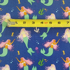 May include: A blue fabric with a repeating pattern of mermaids with pink hair and green tails. The fabric is measured with a yellow tape measure.