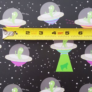 May include: A black and white repeating pattern featuring green aliens in flying saucers with colorful stripes. The text "PEW PEW PEW...", "WHEEEE...", "HIIEEE..." and "000000..." is included in the pattern. A yellow measuring tape with black markings is placed across the pattern.