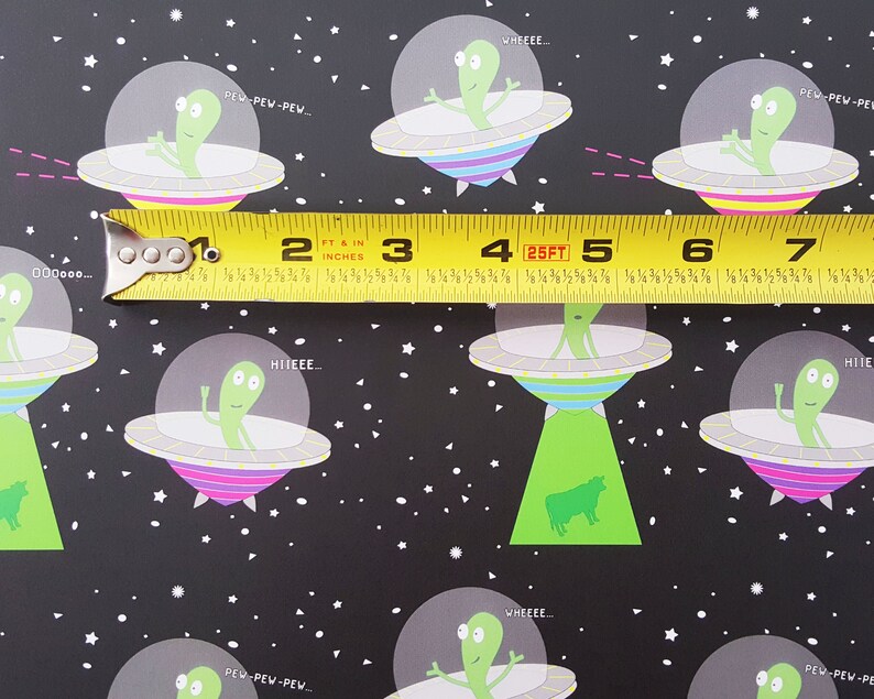 May include: A black and white repeating pattern featuring green aliens in flying saucers with colorful stripes. The text "PEW PEW PEW...", "WHEEEE...", "HIIEEE..." and "000000..." is included in the pattern. A yellow measuring tape with black markings is placed across the pattern.