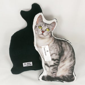 May include: A gray and white tabby cat pillow with green eyes. The pillow is sitting on a black fabric pillow with a white tag that says "Furever Creations".