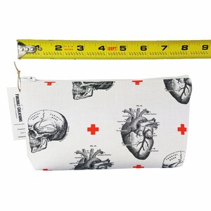 May include: White canvas zippered pouch with a repeating pattern of anatomical heart and skull illustrations in black and white. Red crosses are scattered throughout the pattern. The pouch is approximately 20 cm wide.