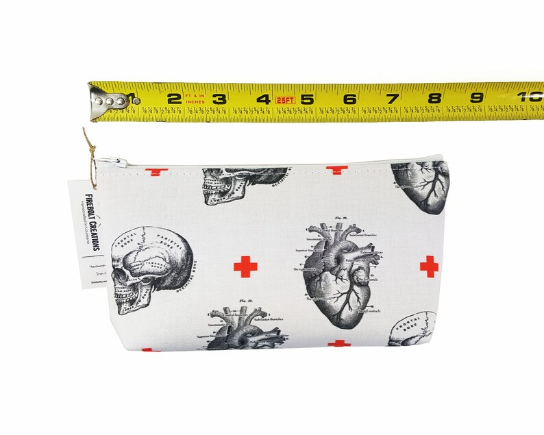 May include: White canvas zippered pouch with a repeating pattern of anatomical heart and skull illustrations in black and white. Red crosses are scattered throughout the pattern. The pouch is approximately 20 cm wide.