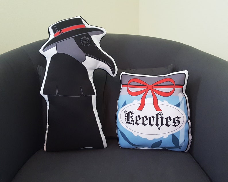 May include: Two decorative cushions, one shaped like a plague doctor with a black hat and robe, and the other shaped like a jar with a red ribbon and the word "Leeeches" on the label.