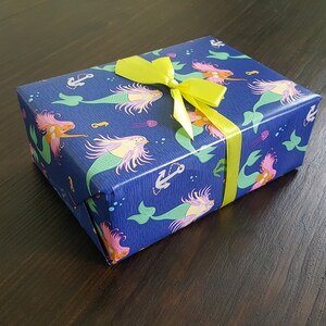 May include: A blue gift box with a yellow bow. The box is wrapped in paper with a mermaid pattern. The mermaids are pink, green, and yellow with long flowing hair. The paper also has anchors and seashells.