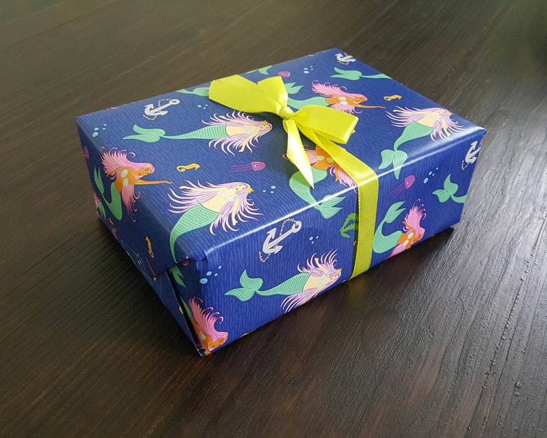 May include: A blue gift box with a yellow bow. The box is wrapped in paper with a mermaid pattern. The mermaids are pink, green, and yellow with long flowing hair. The paper also has anchors and seashells.