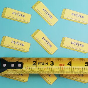 May include: A repeating pattern of yellow butter boxes with the word "BUTTER" in purple on a light blue background. A yellow tape measure is in the foreground, showing measurements in feet and inches.