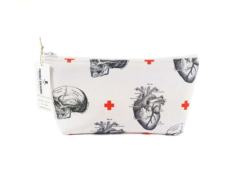 May include: White canvas zippered pouch with a red cross and anatomical illustrations of a heart and skull. The pouch has the text "FREIBOLT CREATIONS" printed on the side.