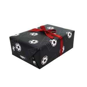Puede incluir: Una caja de regalo negra con una cinta roja. La caja est&aacute; envuelta en papel con un patr&oacute;n de balones de f&uacute;tbol blancos.