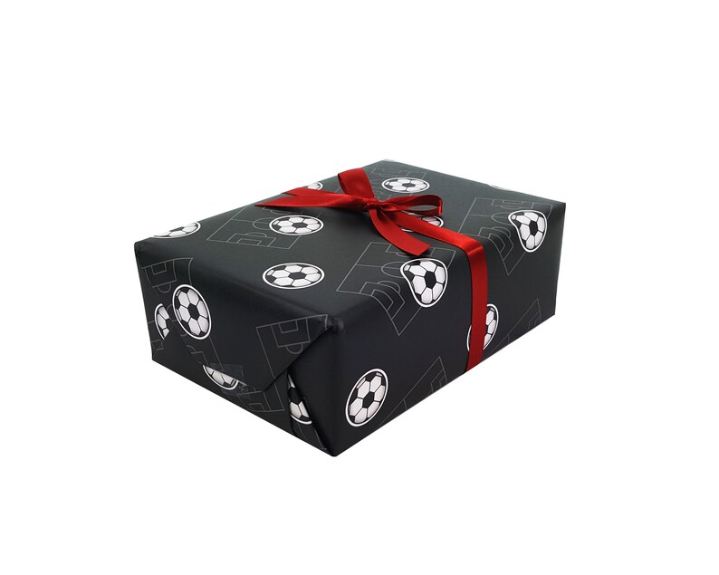 Puede incluir: Una caja de regalo negra con una cinta roja. La caja est&aacute; envuelta en papel con un patr&oacute;n de balones de f&uacute;tbol blancos.