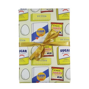 Puede incluir: Papel de regalo amarillo con un patr&oacute;n repetido de ingredientes para hornear, que incluyen mantequilla, harina, az&uacute;car y levadura. El papel est&aacute; atado con una cinta dorada.