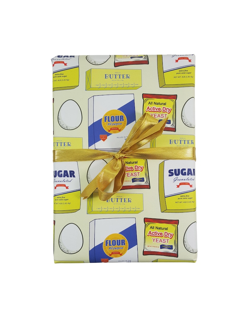 Puede incluir: Papel de regalo amarillo con un patr&oacute;n repetido de ingredientes para hornear, que incluyen mantequilla, harina, az&uacute;car y levadura. El papel est&aacute; atado con una cinta dorada.