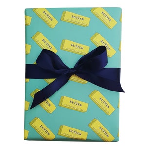 May include: A wrapped gift with teal wrapping paper featuring a repeating pattern of yellow butter sticks with the word "BUTTER" printed on them. A dark blue satin ribbon is tied in a bow around the gift.