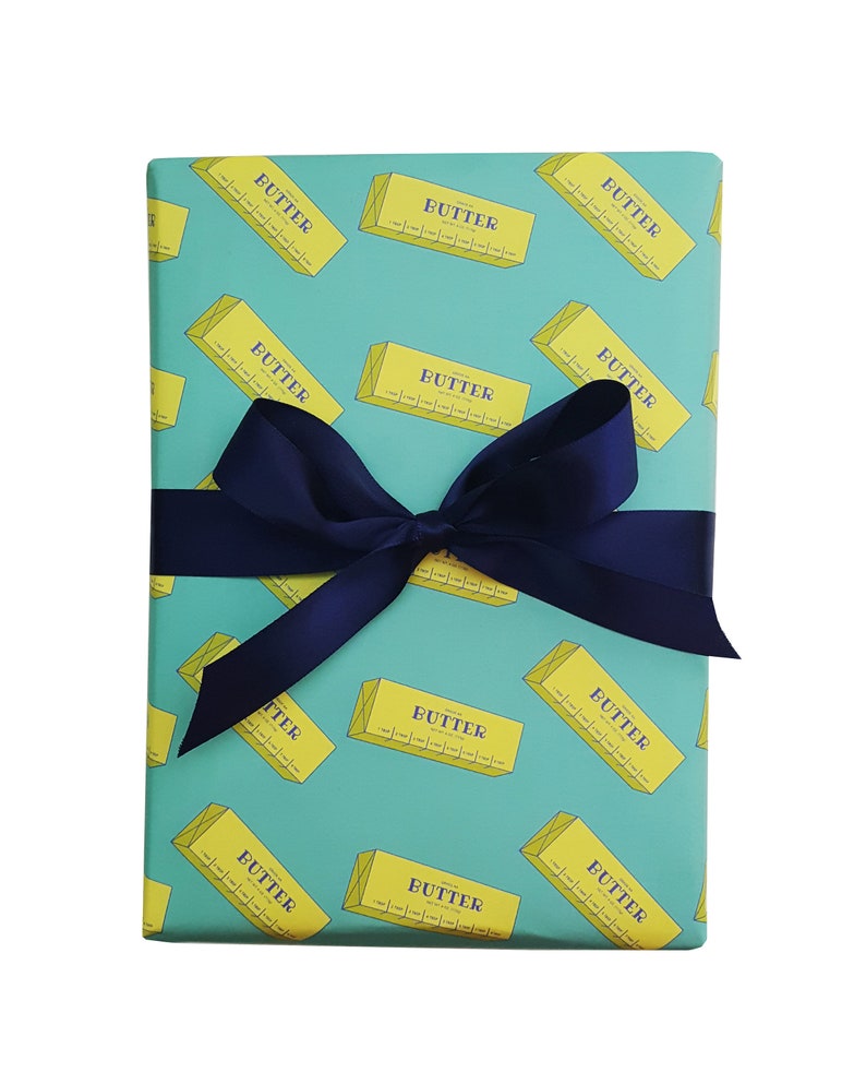 May include: A wrapped gift with teal wrapping paper featuring a repeating pattern of yellow butter sticks with the word "BUTTER" printed on them. A dark blue satin ribbon is tied in a bow around the gift.