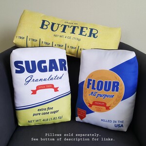 Puede incluir: Tres almohadas decorativas con un dise&ntilde;o vintage de empaque de alimentos. Las almohadas son amarillas, blancas y azules. La almohada amarilla dice "Grade AA Butter" con medidas en onzas. La almohada blanca dice "Sugar Granulated" con medidas en libras. La almohada azul dice "Flour All Purpose" con medidas en libras.