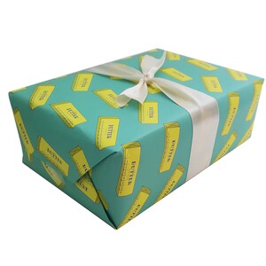 May include: A wrapped gift box with teal wrapping paper featuring a repeating pattern of yellow butter sticks. The box is tied with a white satin ribbon. The word "BUTTER" is printed on the butter sticks.