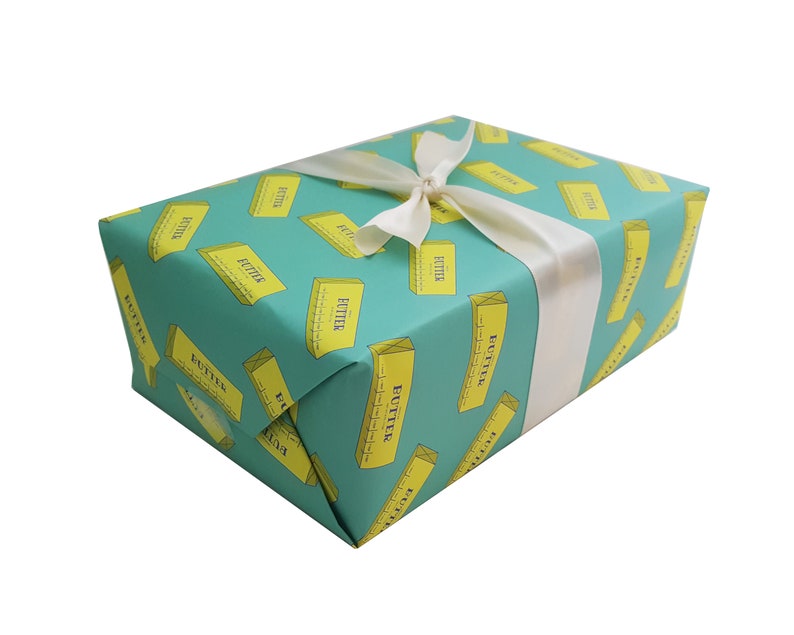May include: A wrapped gift box with teal wrapping paper featuring a repeating pattern of yellow butter sticks. The box is tied with a white satin ribbon. The word "BUTTER" is printed on the butter sticks.