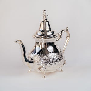 May include: A silver teapot with an ornate design and a lid with a finial. The teapot has a round body and a spout on the side. It sits on three legs.