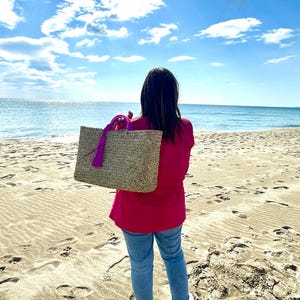 May include: A woven straw tote bag with a purple handle and tassel is held by a person on a sandy beach. The bag is rectangular and appears to be empty. The person is wearing a pink top, blue jeans, and white trainers, with the ocean and sky in the background.
