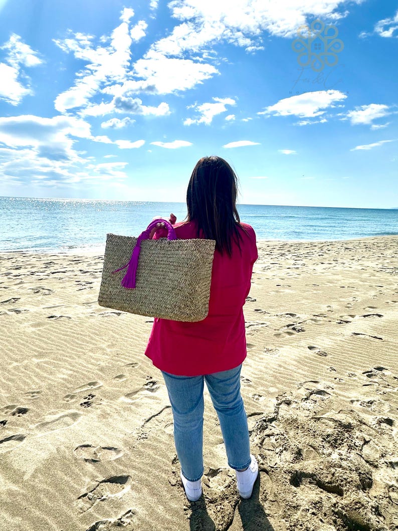 May include: A woven straw tote bag with a purple handle and tassel is held by a person on a sandy beach. The bag is rectangular and appears to be empty. The person is wearing a pink top, blue jeans, and white trainers, with the ocean and sky in the background.