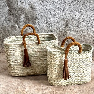 May include: Two rectangular woven straw baskets with brown leather handles and tassel details. The baskets are of different sizes and have a natural colour. They are set against a textured grey background.